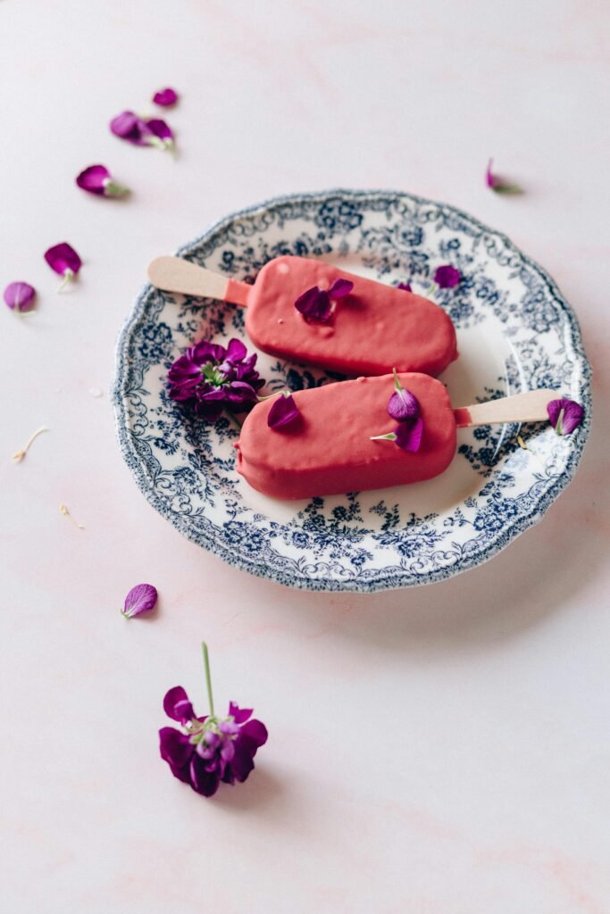 Delicate pink popsicles adorned with flower petals, placed on a decorative blue and white plate on a light surface.