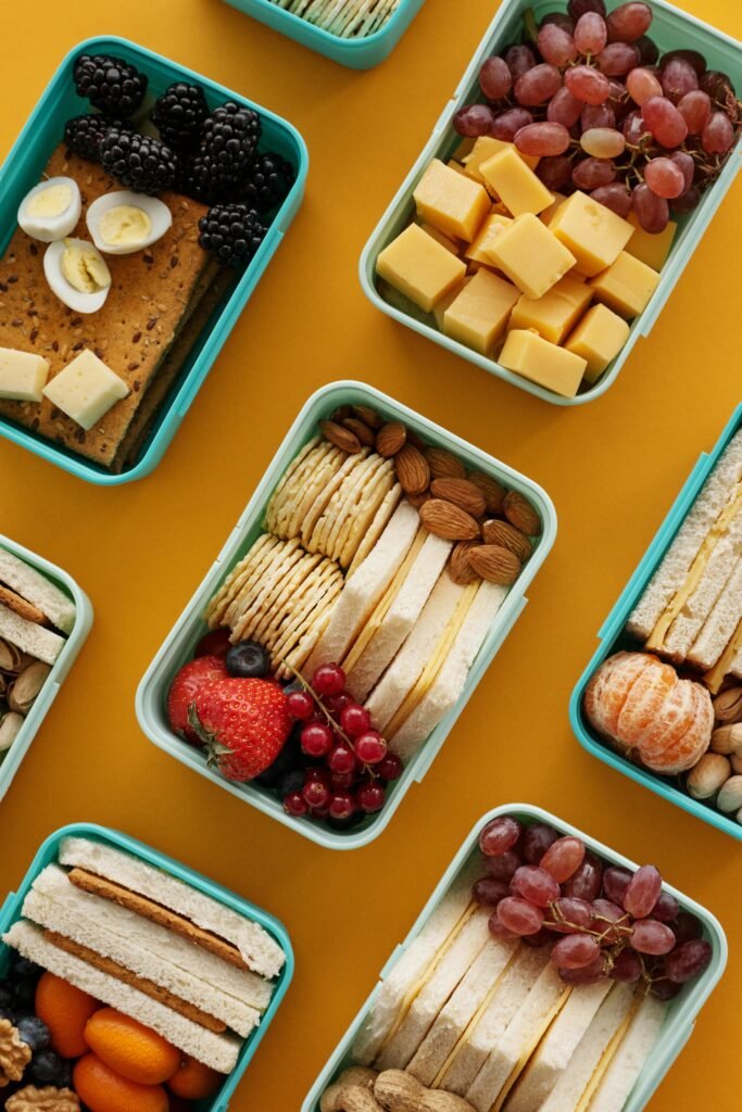 pexels-photo-5971975-5971975 Assorted lunch boxes with snacks, fruits, and cheese on a vibrant yellow background.