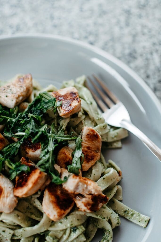 Close-up of a delicious chicken pesto pasta dish featuring grilled chicken, herbs, and a fork.