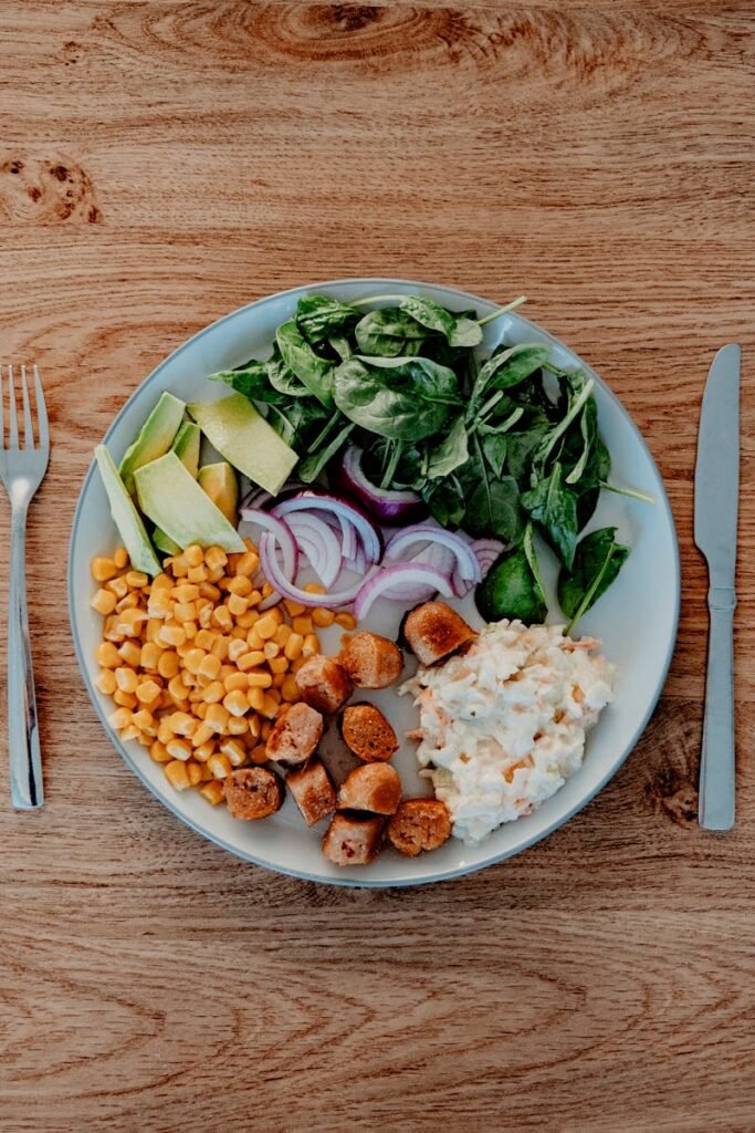 Delicious plate featuring avocado, corn, salad, sausages, and mashed potatoes on a wooden table.